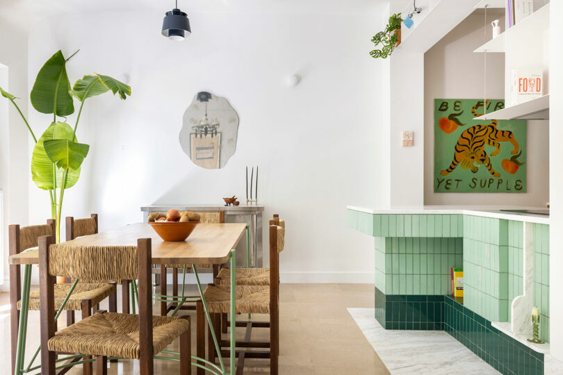 Bright dining area with a wooden table, woven chairs, and a large green plant next to a fireplace.