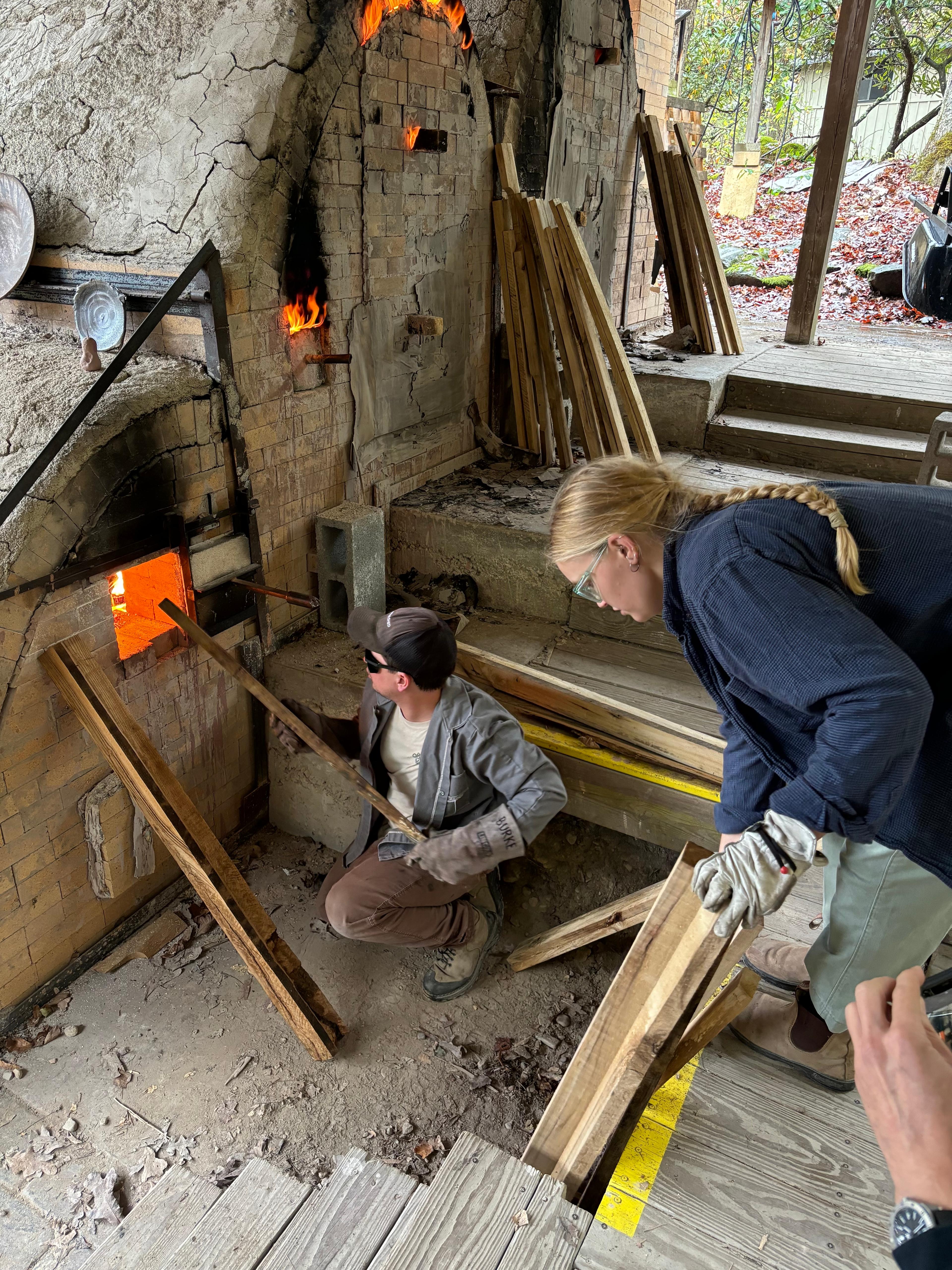 Meghan Burke carefully feeding wood into the intense opening of a glowing ceramic kiln.