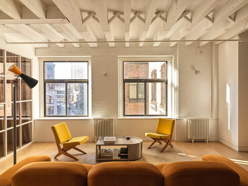 A bright living room with yellow accent chairs and natural light streaming through three large windows.