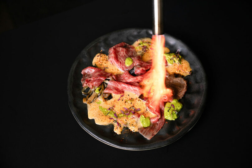 A chef using a torch to char vegetables and meat on a black plate.