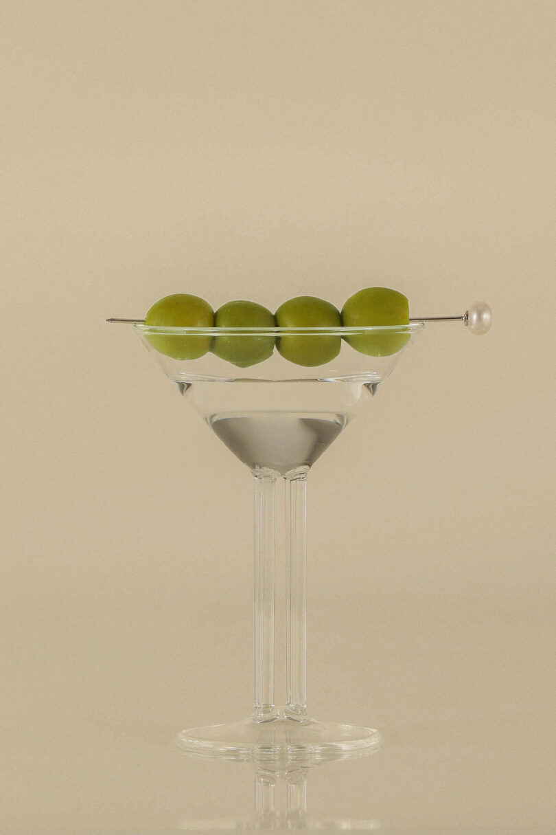 Close-up of a mini martini glass with a four-column stem and four olives on a cocktail pick.