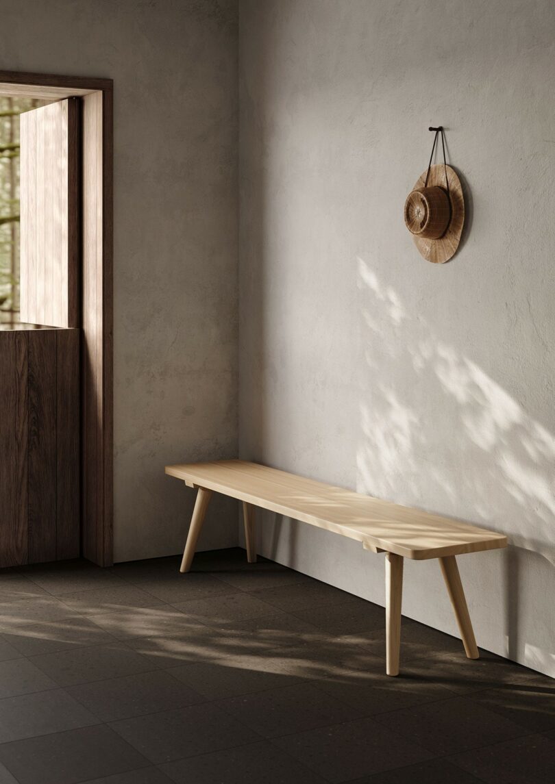A wooden bench against a light wall with a straw hat hanging nearby and sunlight streaming through a door.