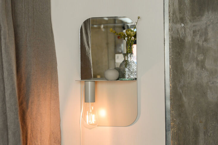 A grey metal wall shelf featuring an integrated circular mirror and lamp.