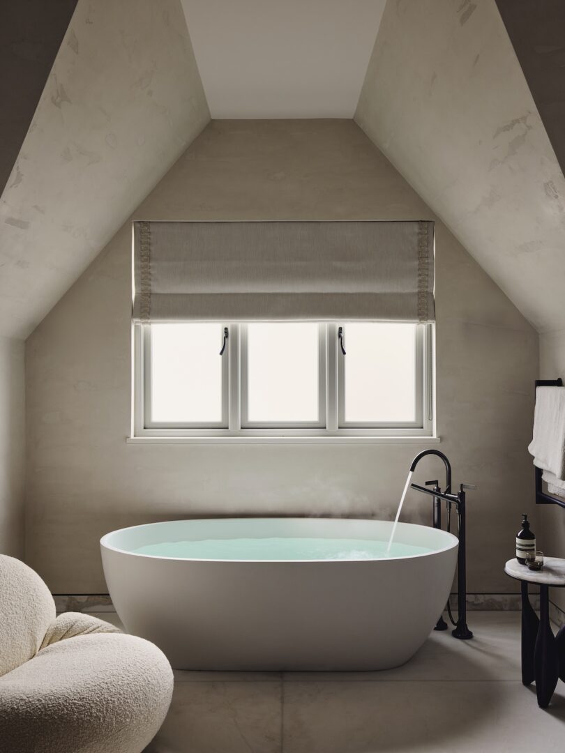 A modern bathroom featuring an oval freestanding bathtub under a window with a neutral Roman shade.
