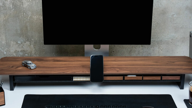 Minimalist desk setup with a Balolo wooden monitor stand and neatly organized accessories against a concrete wall.