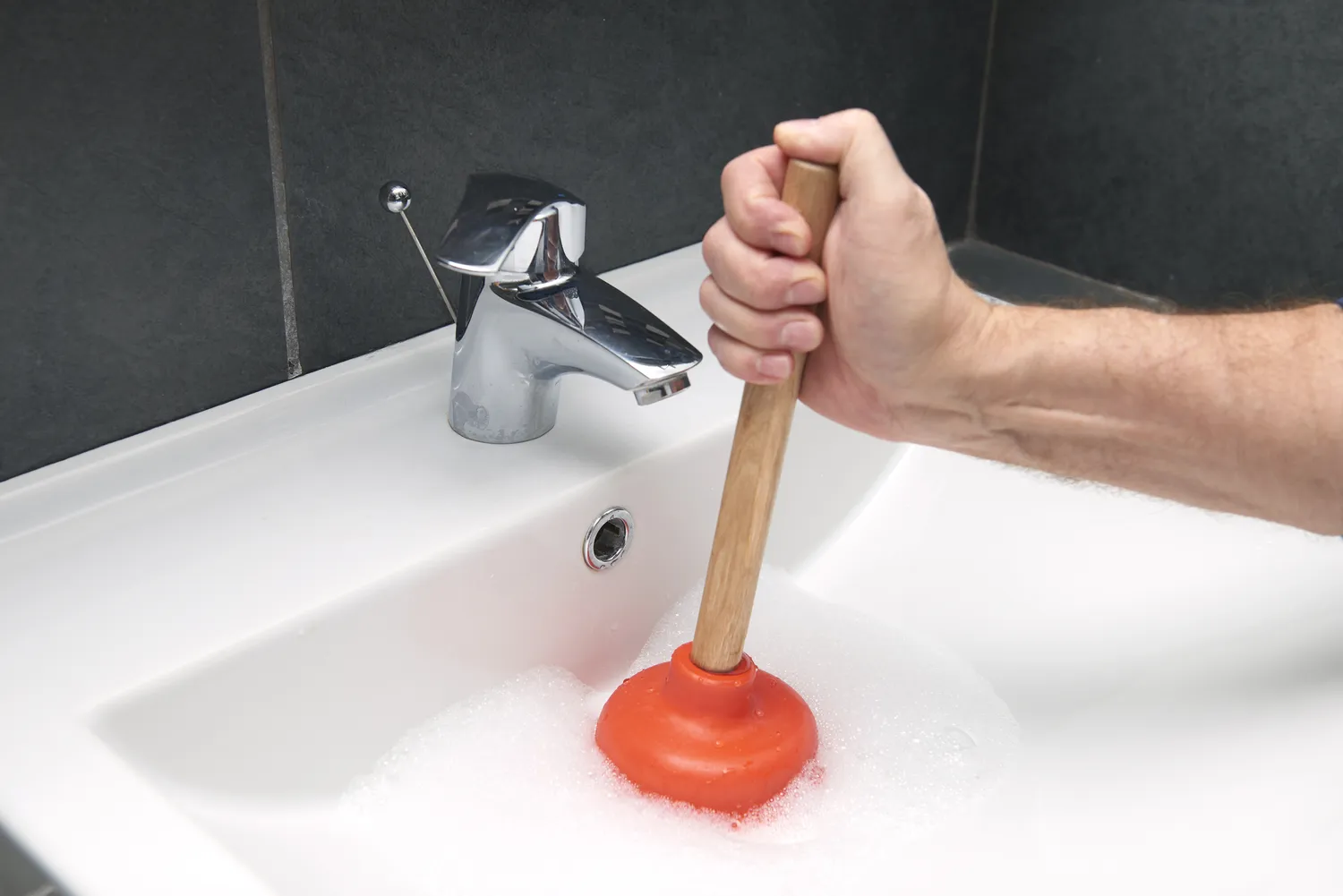 A person using an orange plunger on a modern bathroom sink with soapy water to clear a clog.