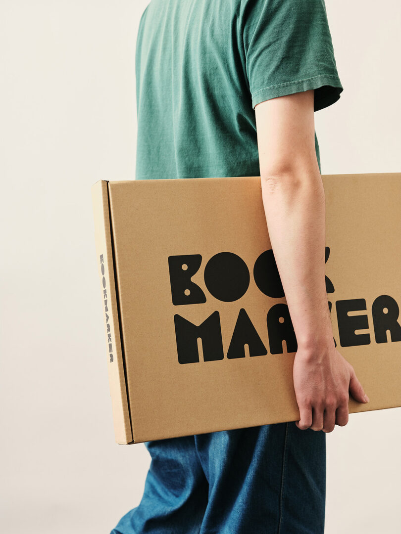 A person holding a slim rectangular cardboard box labeled BOOKMARKER, illustrating the compact flat-pack shipping.