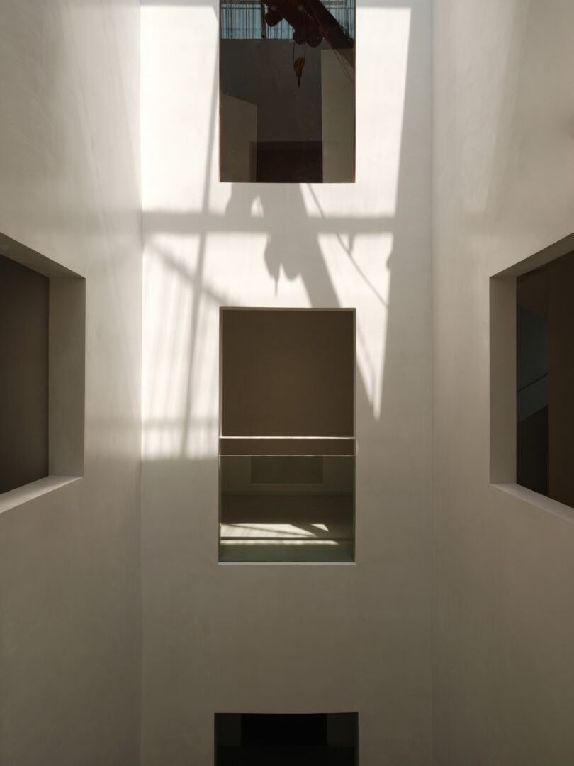 Tall white minimalist interior with light casting sharp shadows through a grid-like ceiling window.