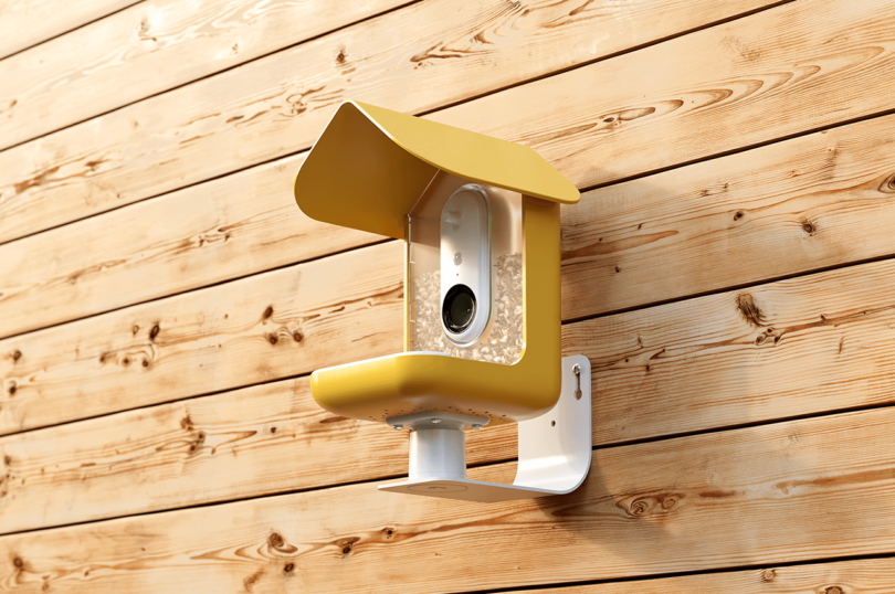 A yellow Bird Buddy smart feeder in high detail showing the camera lens and feeding area.