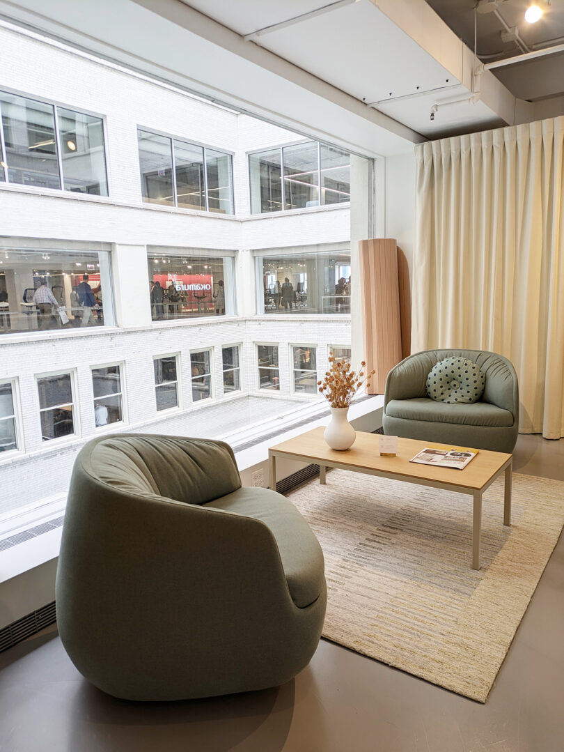 Two sage green armchairs and a light wood table in a sunlit office lounge area near a large window.