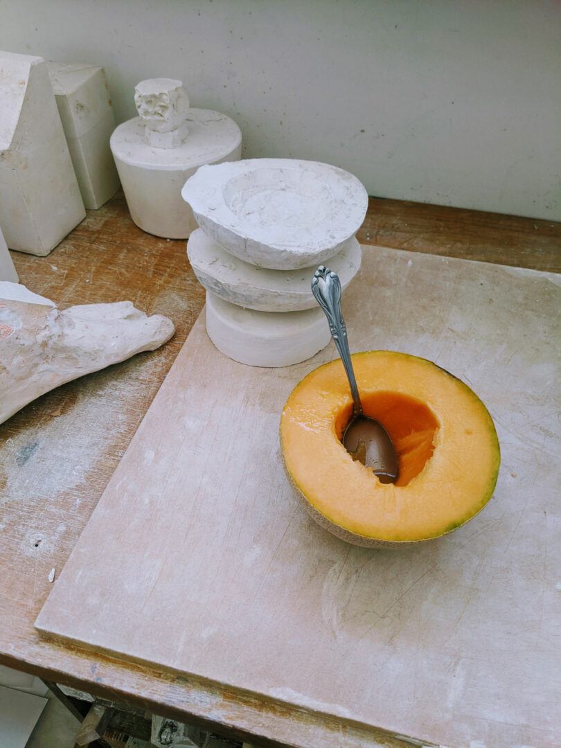 A halved cantaloupe with a spoon next to white plaster sculptural forms in a studio.