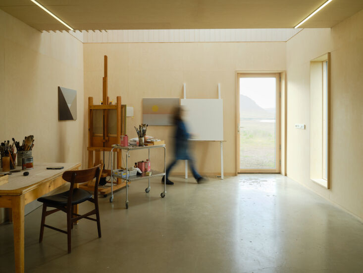 A woman working in a high-ceilinged art studio with white walls and a large window looking out at the landscape.