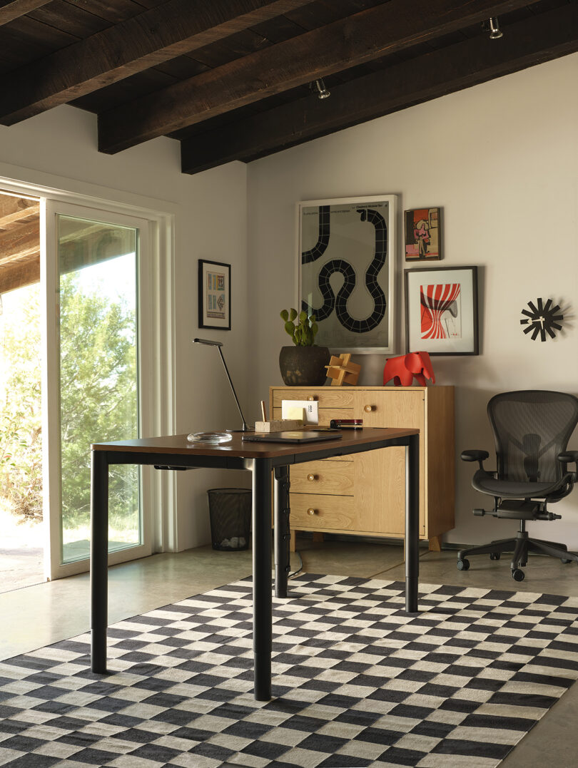 A wooden veneer Spout desk paired with a checkered rug and natural window lighting.