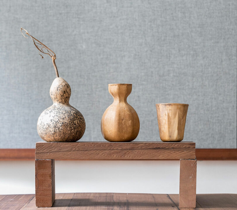 Three handcrafted wooden vases of varying shapes and a dried branch on a rustic wooden bench.