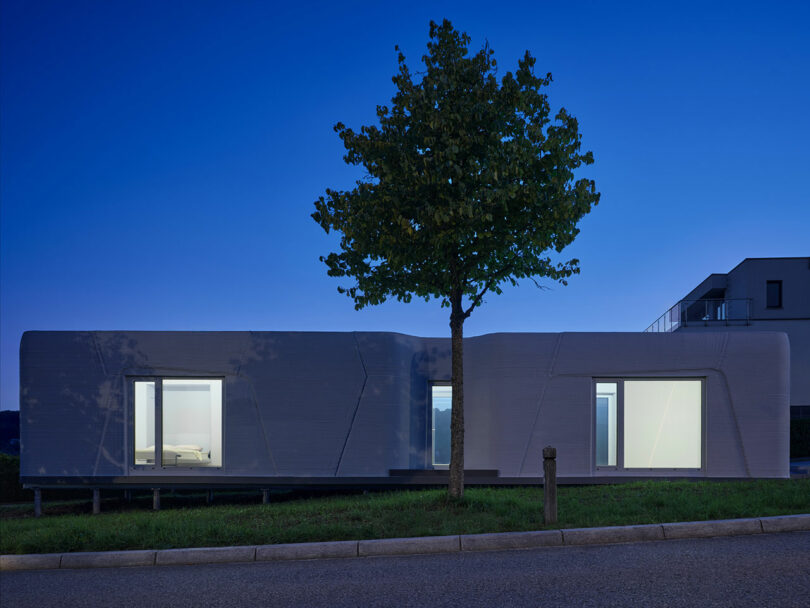 A modern 3D-printed house illuminated from the inside at dusk, showing a warm and inviting living space.