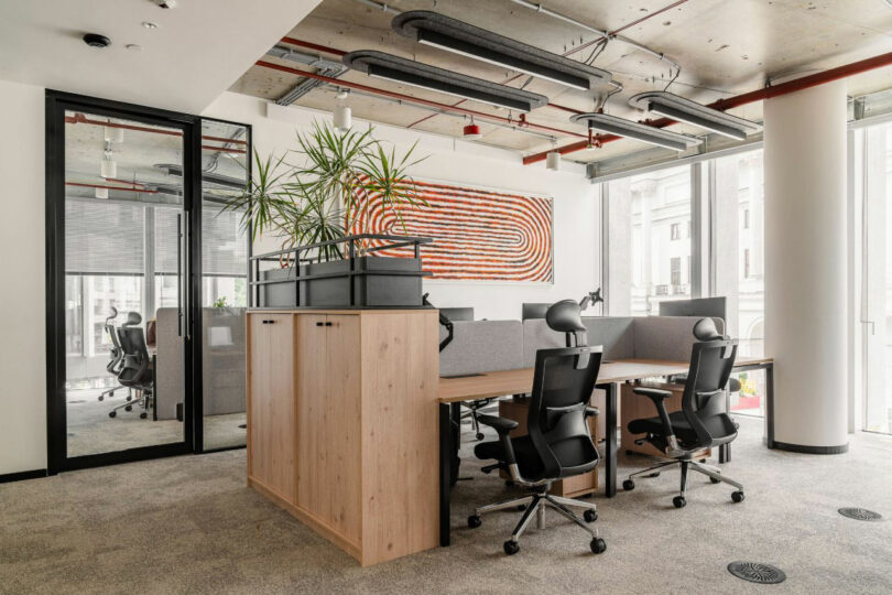 Modern open-plan office with white desks, green plants, and colorful abstract art on the far wall.