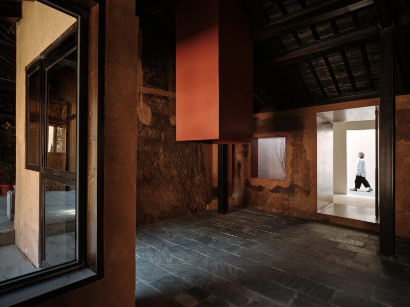 Person walking through a bright doorway in a room with textured earthen walls and a suspended red structure.