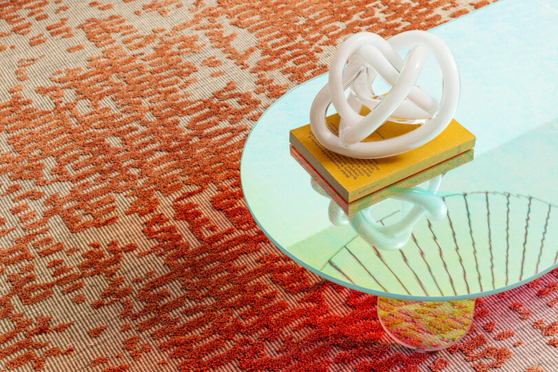 Close-up of a glass coffee table reflecting a white knotted sculpture on a red patterned rug.
