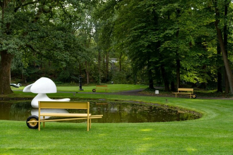 A wide shot of several yellow benches scattered on a lawn near a pond and white sculptures.