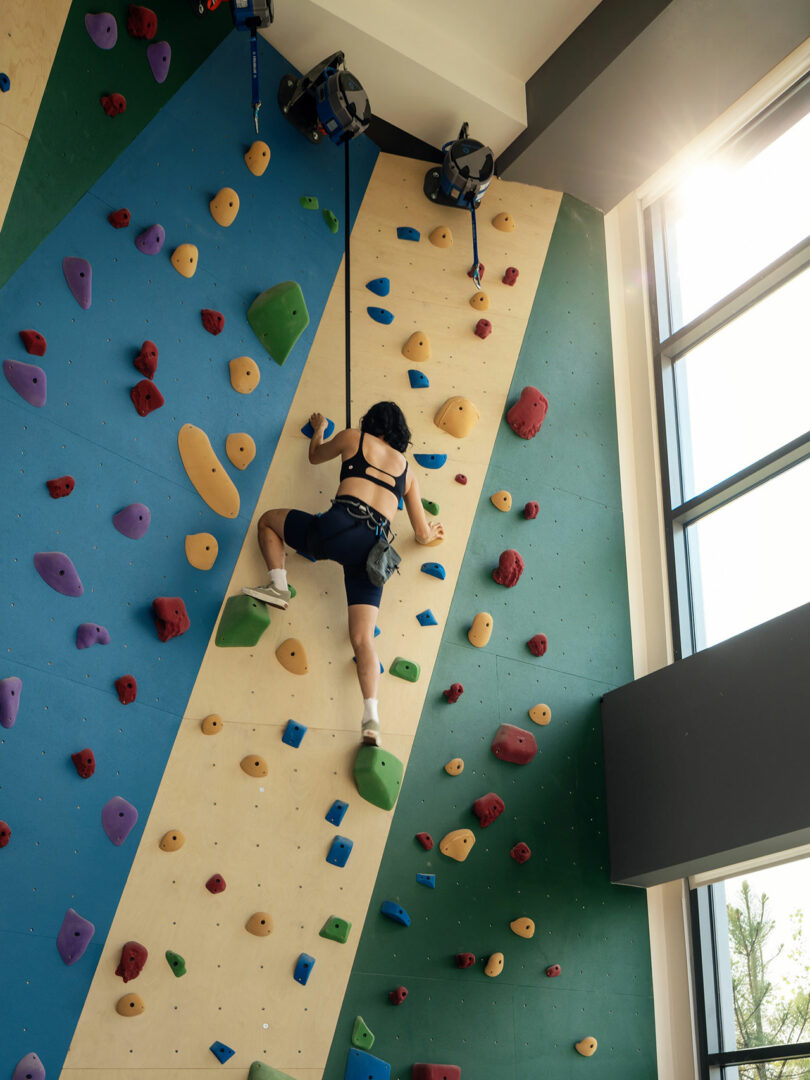 A person climbing an indoor rock wall inside a brightly lit fitness area with large windows.