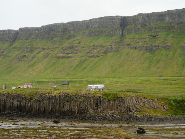 Aerial-style view of a small cluster of farm buildings in a vast, grassy Icelandic landscape near the coast.