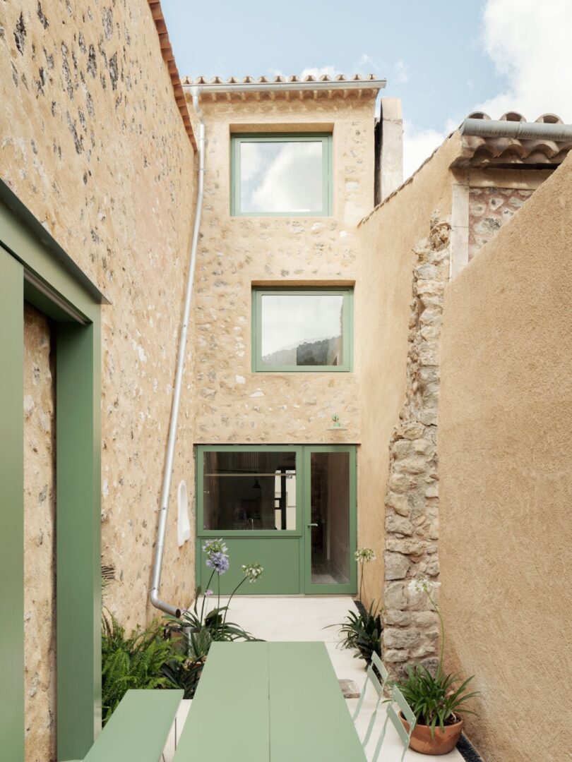 A narrow courtyard with stone walls, a long green outdoor table, and green-framed windows.