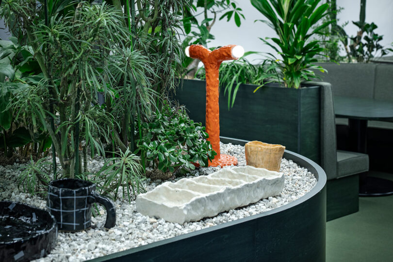 A small indoor garden with white gravel, ceramic objects, and an orange Y-shaped lamp next to a green booth.