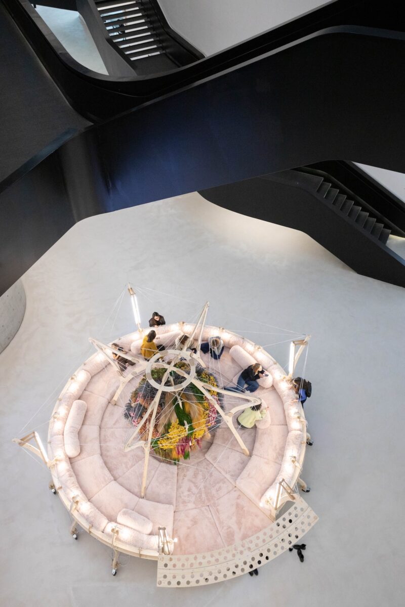 A group of museum-goers sitting on a circular, multi-tiered sofa integrated with plants and lighting.