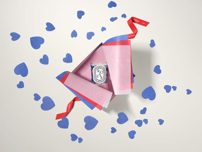 A pink and red gift box featuring a scented candle surrounded by blue heart cutouts and a red ribbon.