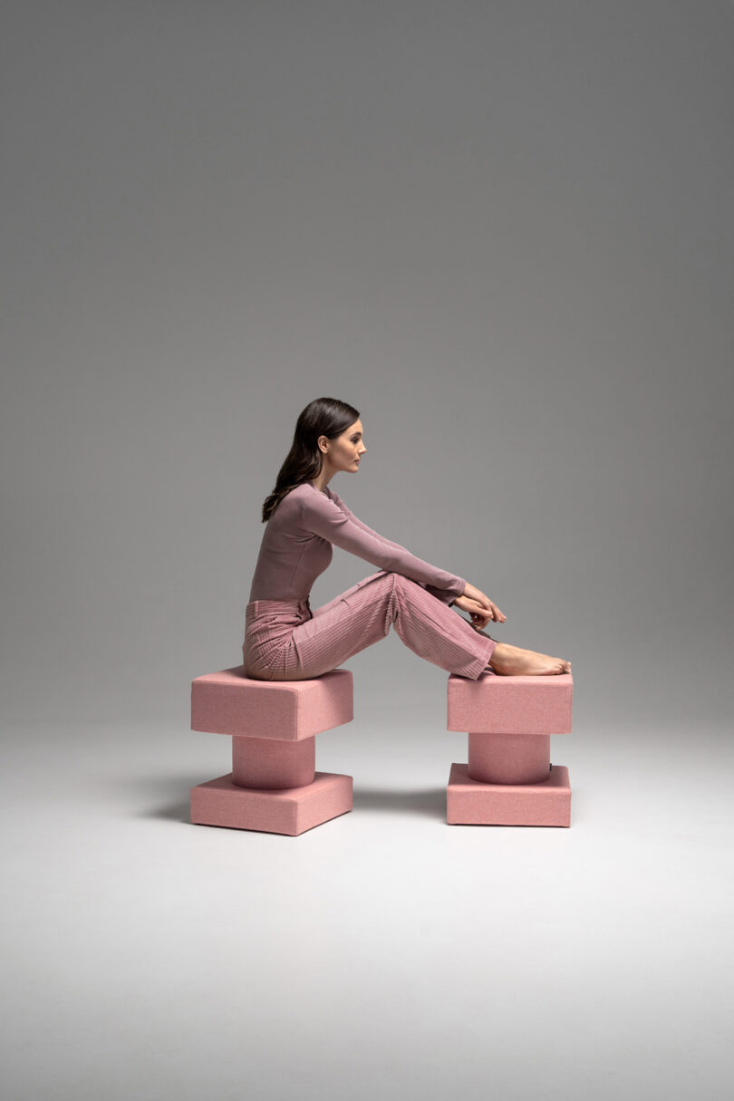 A woman sits comfortably on two stacked pink geometric blocks in a minimalist studio.