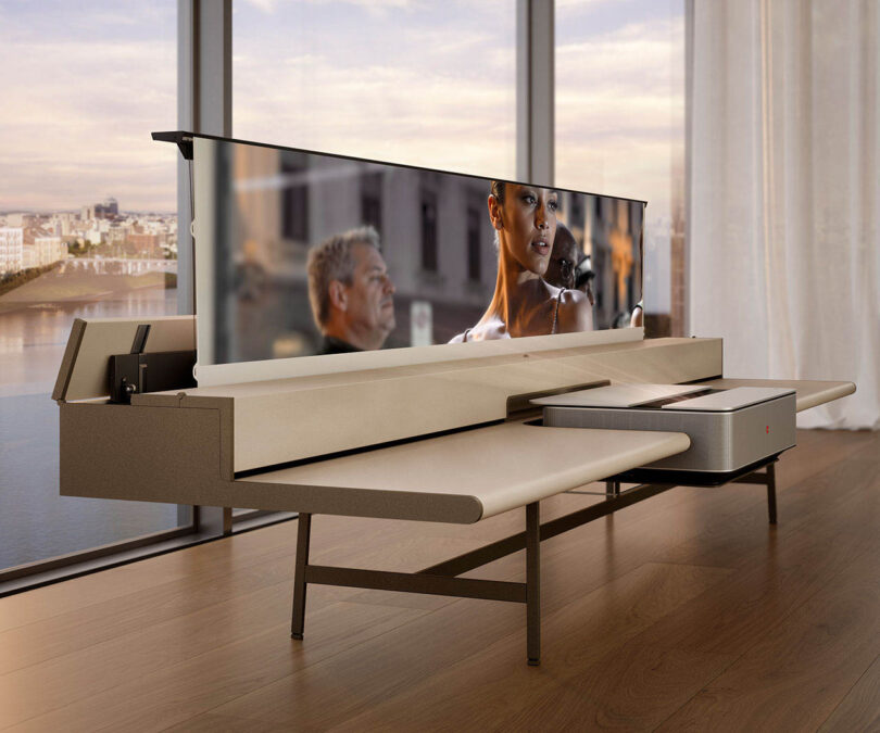 A partially raised retractable screen emerging from a minimalist beige sideboard in front of a window.