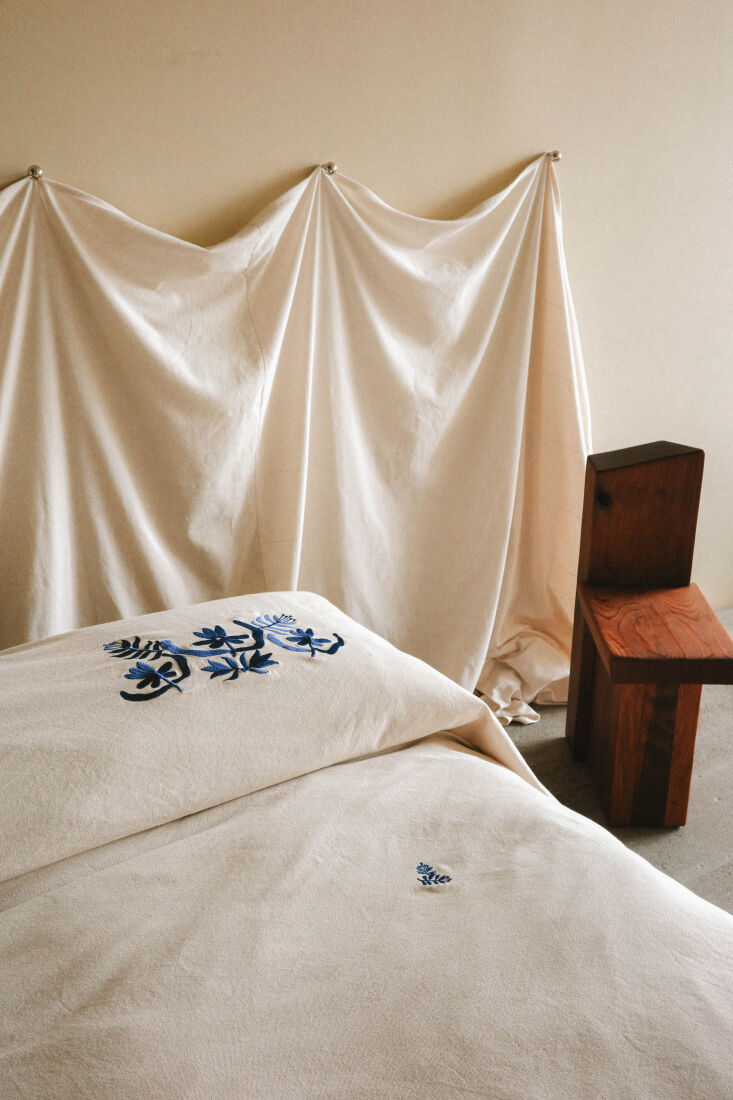 Embroidered Marta Coverlet in Natural and Azure pinned to the wall behind a bed.