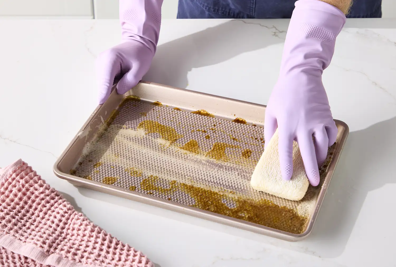 Hands in lavender gloves scrubbing a stained metal baking pan with a sponge.
