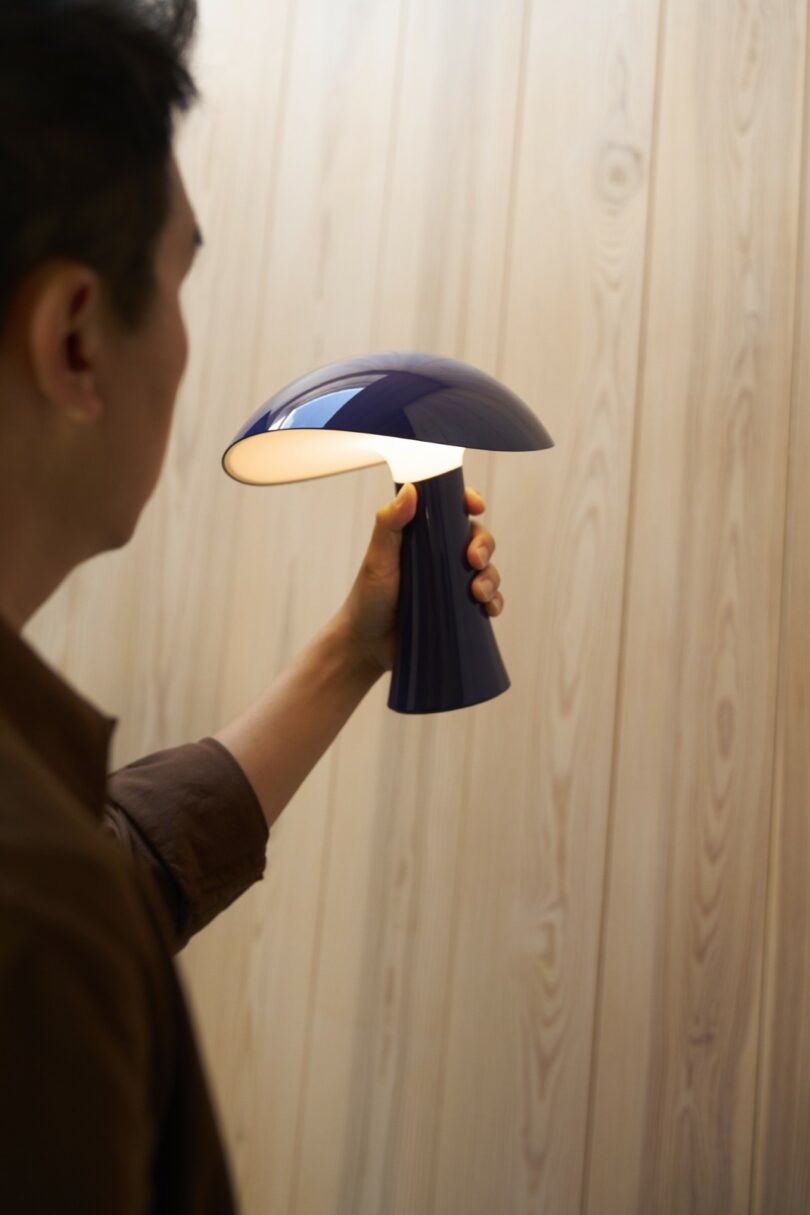 A person holding a lit dark blue mushroom-shaped lamp against a wooden wall.