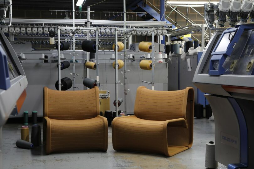 Two orange Isomi Knit One chairs on a factory floor with knitting machines and spools in the background.