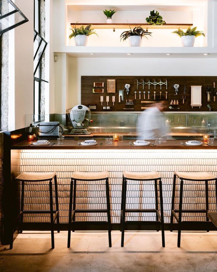 A pasta-making counter where kitchen tools are hung on the wall behind chefs and bar seating.