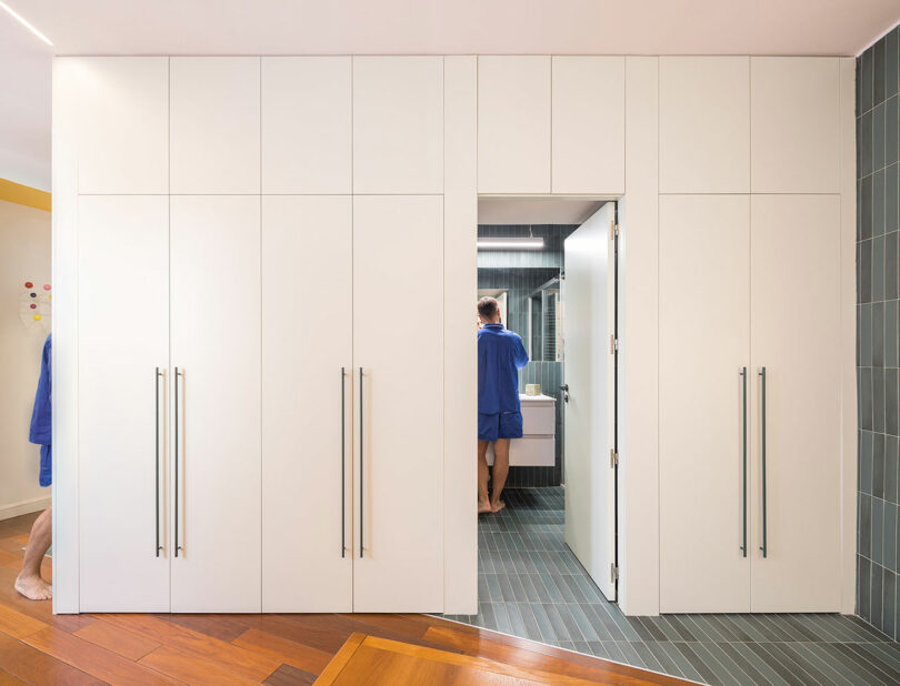 View through a white storage cabinet door into a bathroom with a person at the sink.