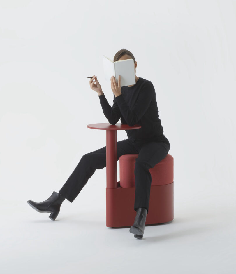 Person in black sitting on a red pony stool while writing in a notebook.