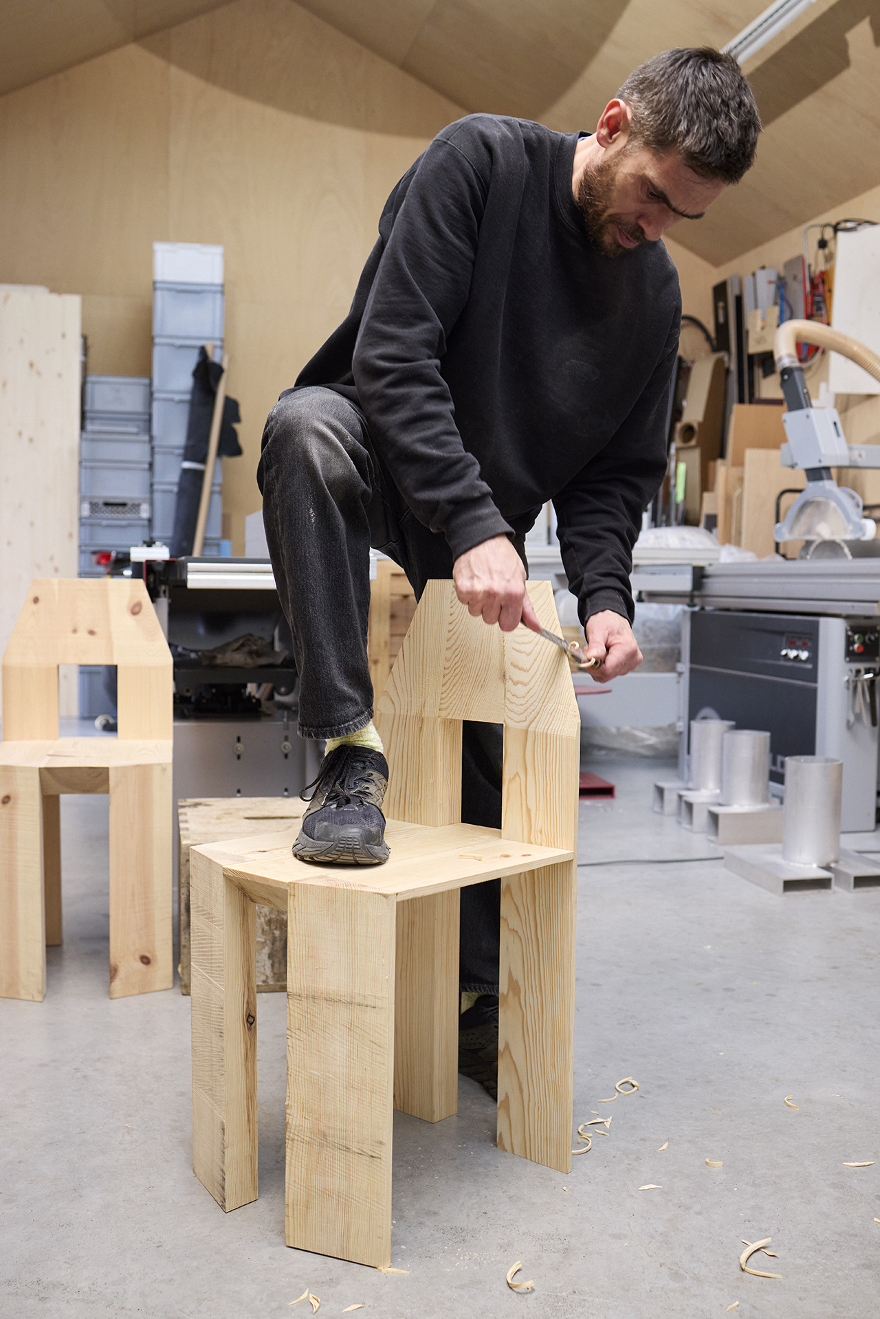 Max Lamb using hand tools to shape a wooden chair in his workshop among wood shavings.
