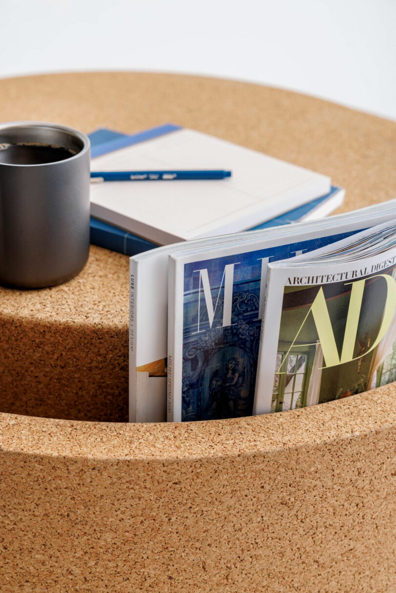A round cork table with a built-in slot used for storing magazines alongside a mug and notebooks.