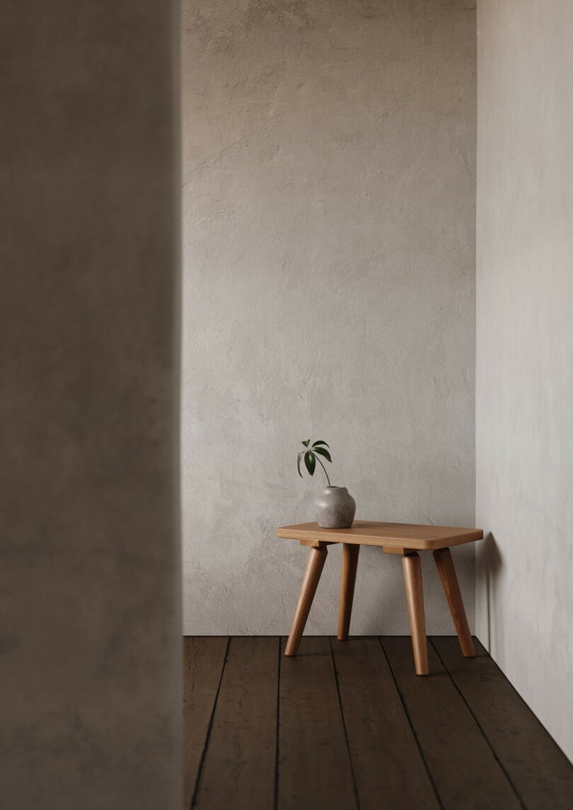 A minimalist four-legged wooden table against a textured beige wall on a dark wood floor.