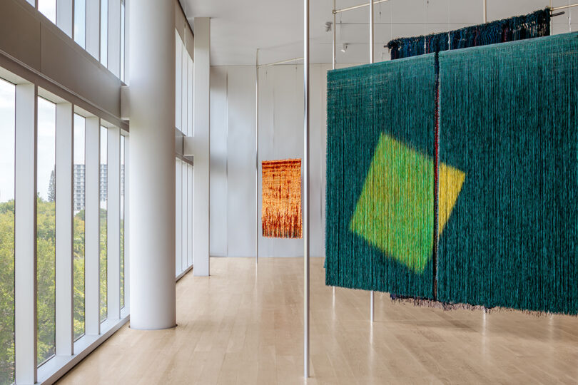 Three large textile installations hanging near floor-to-ceiling windows in a bright gallery.