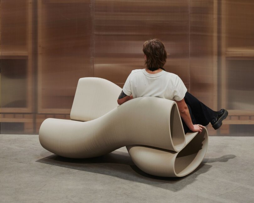 A person sitting on a beige, wave-like sculptural bench with a distinct 3D-printed texture.