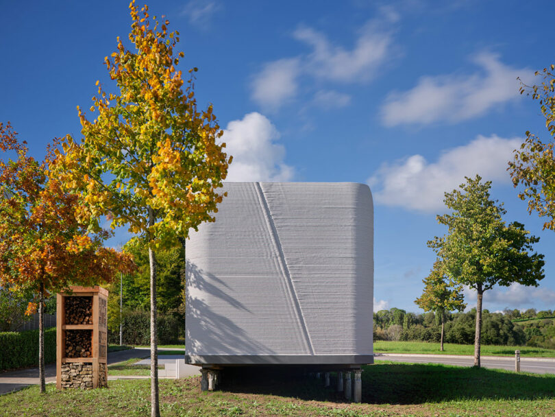 A small, modern 3D-printed home elevated on short stilts in a grassy, wooded area.