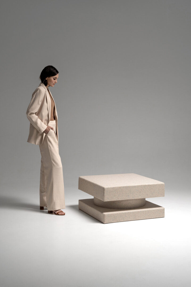 A woman in a beige suit stands next to a modern, square, layered beige TAITŌ table in a minimalist studio.