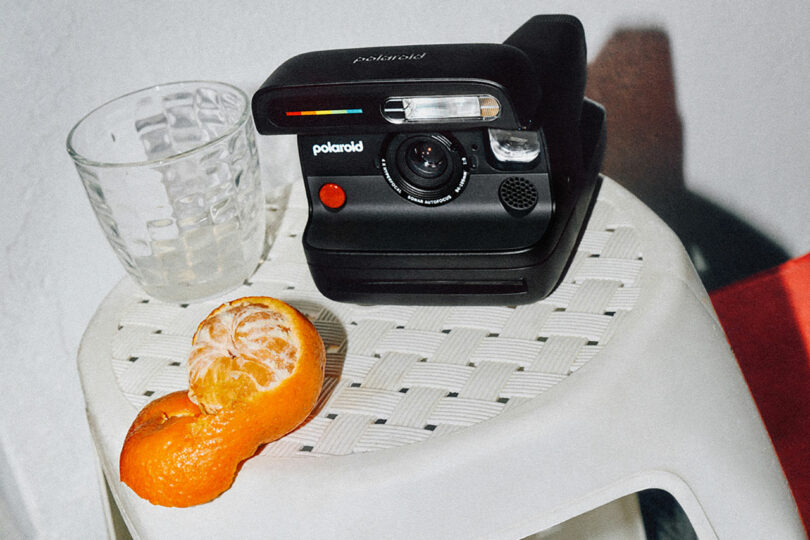 A black Polaroid camera, a glass of water, and a partially peeled orange on a white surface.