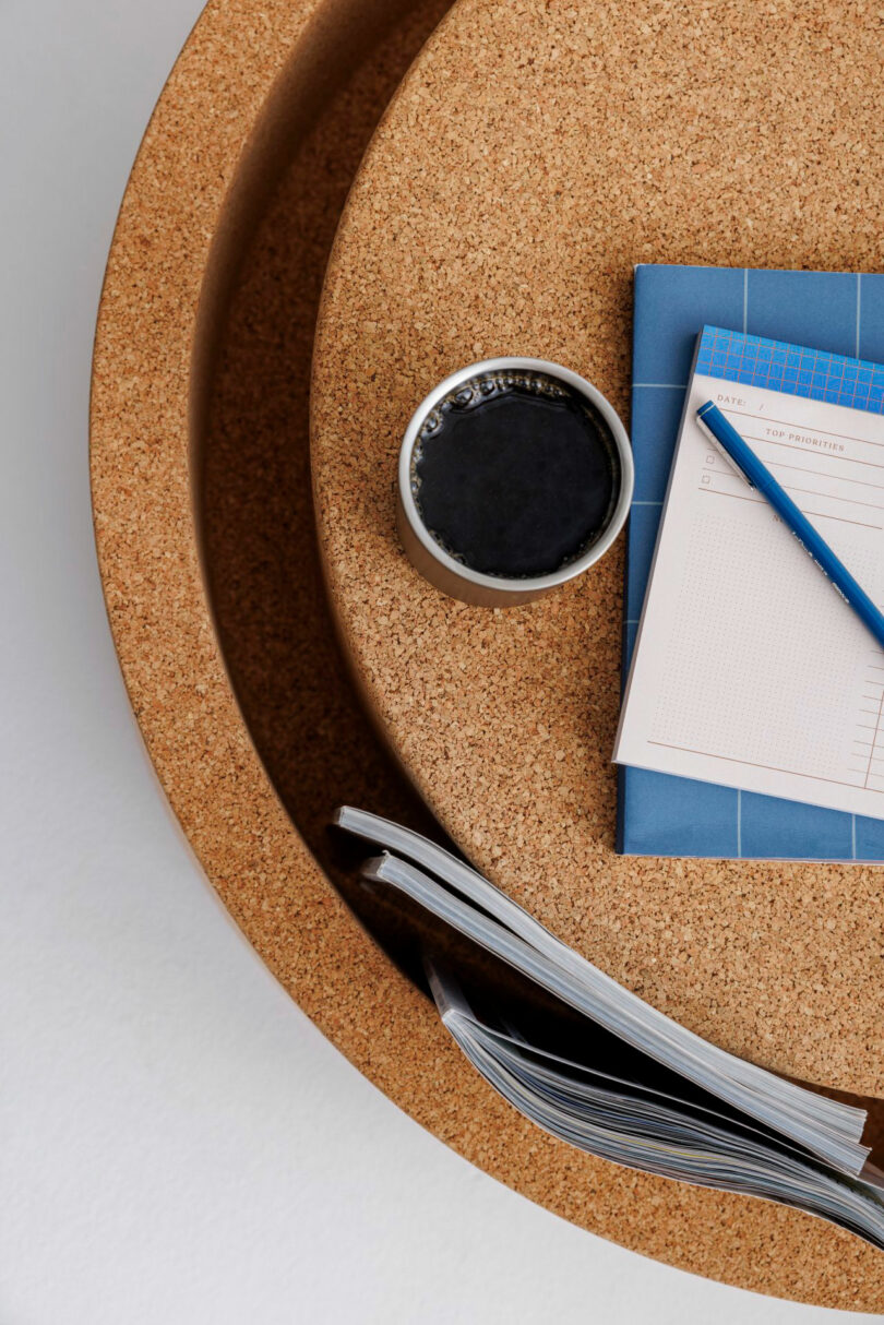 A flat lay perspective of a cork table surface with coffee, a notebook, a pencil, and an open magazine.