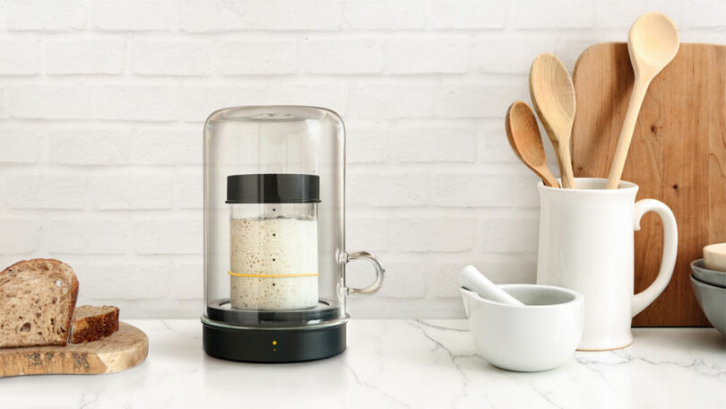 A fermentation jar and fresh loaf of bread on a kitchen counter with wooden tools.