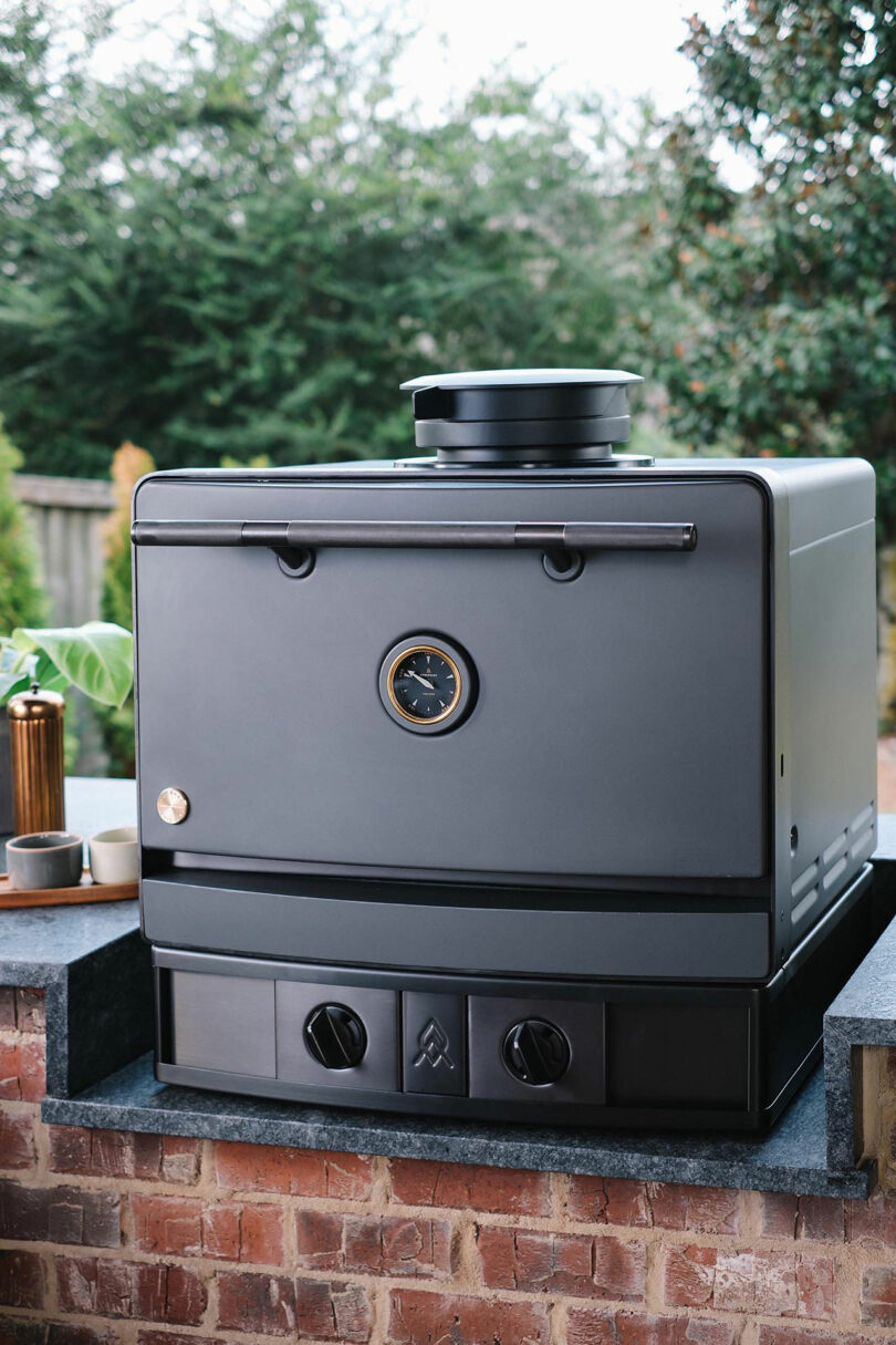 A black Emberhaus charcoal oven on a brick countertop, positioned for backyard pizza making.