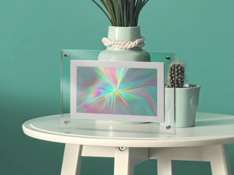 A holographic print in an acrylic frame on a desk with a plant and teal wall background.